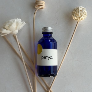 May include: A blue glass bottle with a silver cap and a white label with the word "pêtya." is surrounded by decorative wooden sticks and a white flower. A yellow circle is on the bottle. The arrangement is on a light gray surface.