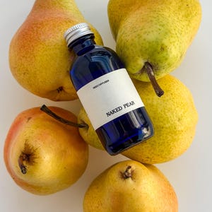May include: A small cobalt blue glass bottle with a silver screw cap and a white label reading REED DIFFUSER NAKED PEAR, surrounded by five yellow and green pears with brown stems. The bottle is positioned centrally among the fresh fruit.