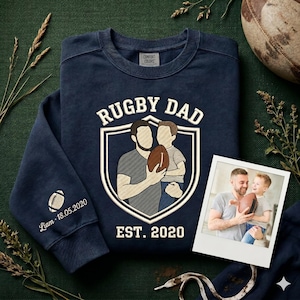 Può includere: Felpa blu navy con la scritta "RUGBY DAD" e un'illustrazione di un padre e un bambino che tengono un pallone da rugby. La manica ha una grafica di pallone da rugby e la scritta "Liam - 18.05.2020". È inclusa anche una foto di un padre e un bambino.