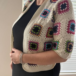 May include: Cream-colored crochet cardigan with colorful square patterns. The squares feature a variety of colors including black, pink, blue, green, and purple. The cardigan has short sleeves and a simple, open front design.