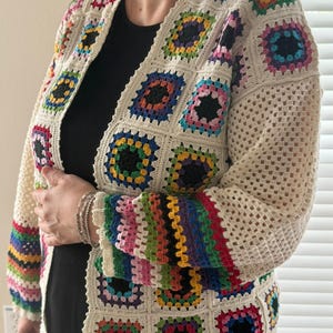 May include: Cream-colored crochet cardigan with colorful square patterns. The squares feature a variety of colors including red, blue, green, and yellow. The sleeves have a rainbow stripe design. The cardigan is open-fronted and appears to be handmade.