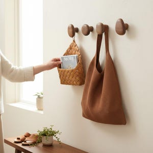 Może przedstawiać: Drewniane haczyki ścienne z plecionym koszem i brązową torbą na kremowej ścianie. Ręka sięga po kosz. Kosz zawiera magazyn ze słowem "MORE" na okładce.