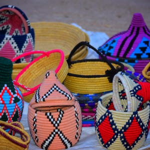 Pode incluir: Uma coleção de cestos e recipientes tecidos coloridos. Os cestos apresentam padrões geométricos em rosa, azul, preto e branco. Alguns têm tampas e alças, enquanto outros são tigelas abertas. A estética geral é artesanal.
