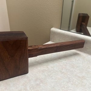 May include: A handcrafted wooden mallet with a dark brown finish. The mallet has a rectangular head and a long, rectangular handle. The wood grain is visible, adding to the rustic aesthetic. The tool is designed for striking or shaping materials.