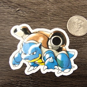 May include: A die-cut sticker of a blue and brown creature with two large cannons on its back, positioned next to a silver United States quarter coin for scale, featuring a white border around the character's illustration.