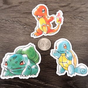May include: Three die-cut stickers featuring cartoon creatures: an orange fire-type, a green plant-type, and a light blue water-type. A silver US quarter coin is placed in the center for scale, measuring approximately 0.955 inches in diameter.