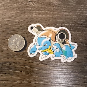 May include: A sticker featuring a blue and brown cartoon turtle-like creature with two cannons on its back. The sticker is next to a U.S. quarter for size comparison. The creature has a stern expression and is outlined in black.