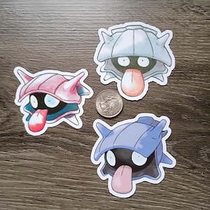 May include: Three die-cut stickers featuring a cartoon character with a shell-like head, a black body, and a tongue sticking out. The stickers are in pink, blue, and gray, with a quarter for size comparison. The stickers are on a wooden surface.