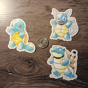 May include: Three colorful stickers featuring Pokémon characters. The stickers depict Squirtle, Wartortle, and Blastoise, each in a unique pose. A U.S. quarter is included for size reference. The stickers are on a dark wood grain surface.