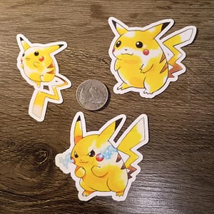 May include: Three yellow Pikachu stickers with black and white accents. One sticker shows Pikachu with a happy expression, another with a neutral expression, and the third with an angry expression and electric sparks. A quarter coin is next to the stickers.