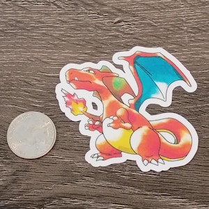May include: A colorful sticker featuring a cartoon dragon with orange, red, yellow, and blue accents. The dragon is holding a flame in its hand. A quarter is next to the sticker for size comparison.