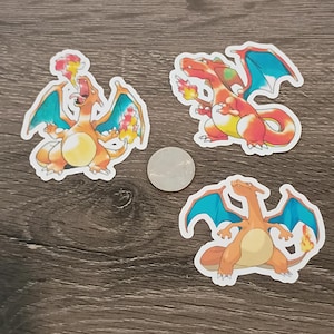 May include: Three orange and blue Charizard stickers are displayed on a wooden surface. The stickers feature the popular Pokémon character in various poses, with flames and wings. A silver coin is also present.