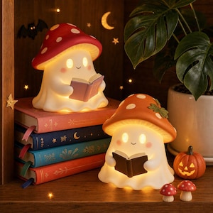 May include: Two illuminated ghost figurines with mushroom caps, one red and one orange, each holding a small open book. The ghosts sit among a stack of four colorful hardcover books, a small jack-o'-lantern, and two tiny mushroom ornaments.