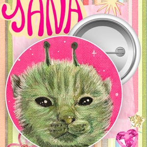 May include: A circular pin-back button featuring a green cat-like creature with two antennae on a bright pink background. The text STUDIO YANA is printed in large pink letters above the button. The back of the pin shows a silver metal mechanism.
