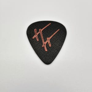 May include: A black guitar pick with a textured surface and a coral-colored graphic design. The pick has a teardrop shape and is designed for use with a guitar. The graphic appears to be a stylized letter or symbol.