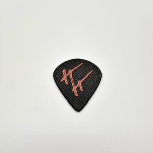 May include: A black guitar pick with a textured surface and a copper-colored design. The design appears to be stylized initials or a logo. The pick is in a teardrop shape.