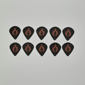 May include: Ten black guitar picks with a flame and leaf design in orange. The picks are arranged in two rows of five against a white background. The picks are teardrop-shaped.