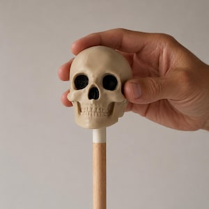 Può includere: Un teschio in plastica beige fissato su un bastoncino di legno chiaro. Il teschio presenta orbite profonde e scure e una cavità nasale dettagliata, con una finitura liscia e opaca. L'asta di legno è cilindrica e di colore marrone chiaro.