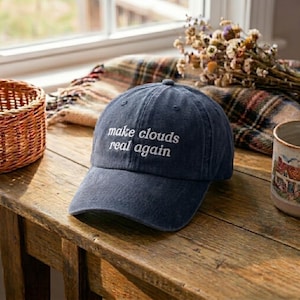 May include: A navy blue baseball cap with the phrase "make clouds real again" embroidered in white. The hat is sitting on a wooden surface, with a basket, a plaid blanket, and a mug in the background.
