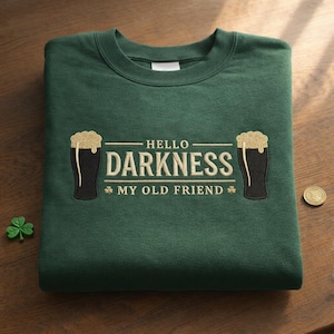 May include: A dark green sweatshirt with the embroidered phrase "HELLO DARKNESS MY OLD FRIEND" and two beer glasses. A small shamrock and a coin are on the wooden surface.