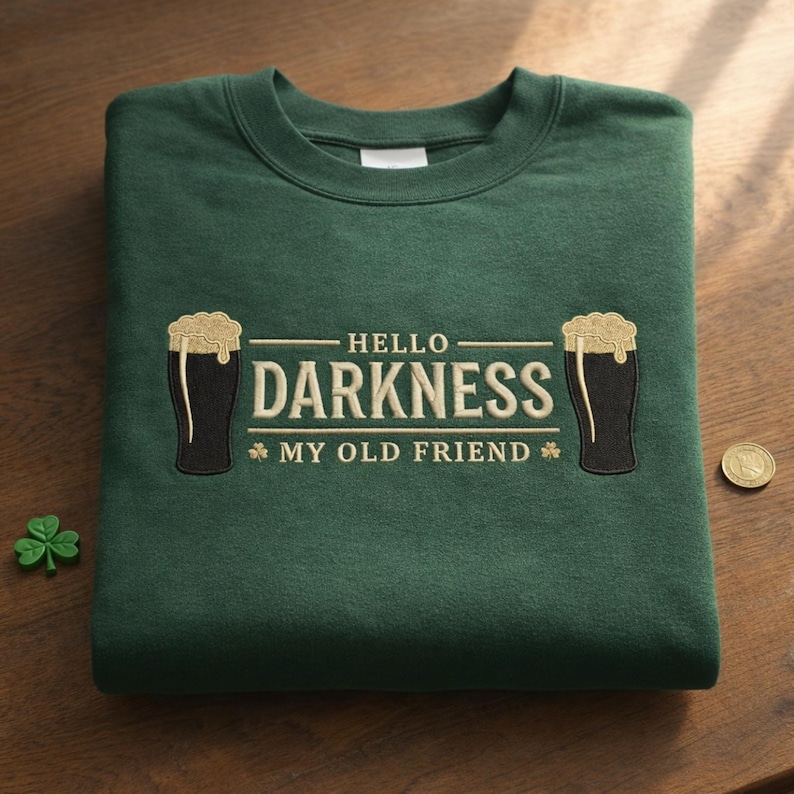 May include: A dark green sweatshirt with the embroidered phrase "HELLO DARKNESS MY OLD FRIEND" and two beer glasses. A small shamrock and a coin are on the wooden surface.