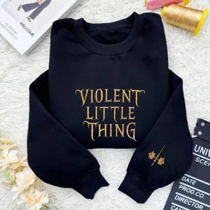 May include: Black sweatshirt with the embroidered text "VIOLENT LITTLE THING" in gold. A gold emblem is on the left sleeve. The sweatshirt is displayed on a white, fluffy surface.