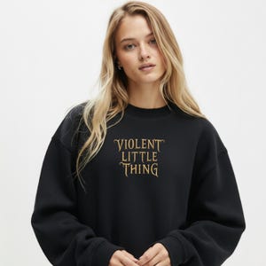 May include: Black crew neck sweatshirt with the text "VIOLENT LITTLE THING" in gold lettering. The sweatshirt is a solid black color and has long sleeves. The overall style is casual and comfortable.
