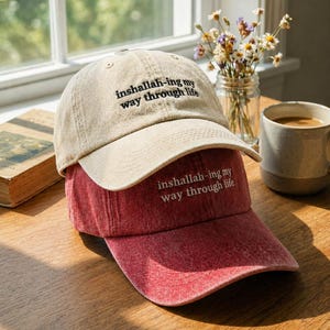 May include: Two baseball caps, one tan and one red, are stacked on a wooden surface. The tan cap has black embroidered text that reads "inshallah-ing my way through life." The red cap has the same text embroidered in white.