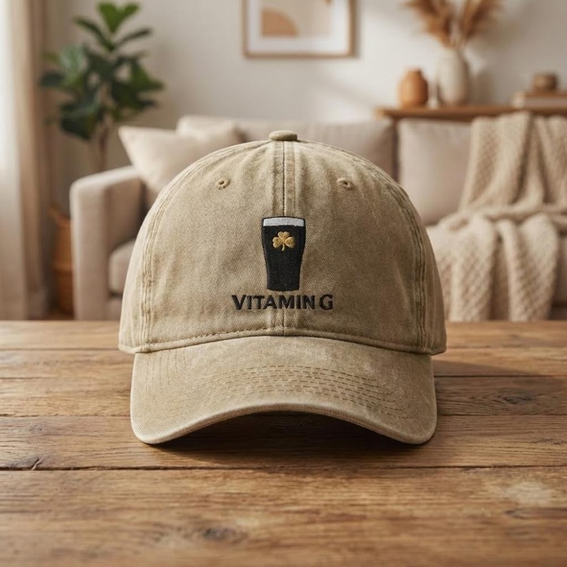 K&ouml;nnte beinhalten: Beige Baseballkappe mit einer schwarzen Grafik eines Pints Stout mit einem Kleeblatt und dem Text "VITAMIN G" auf der Vorderseite gestickt. Die Kappe liegt auf einer Holzoberfl&auml;che.