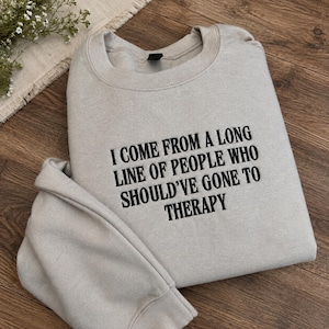 May include: A light gray crewneck sweatshirt featuring black embroidered text that reads, I COME FROM A LONG LINE OF PEOPLE WHO SHOULD'VE GONE TO THERAPY. The garment has long sleeves, a ribbed collar, and a ribbed hem, showcasing a clean design.