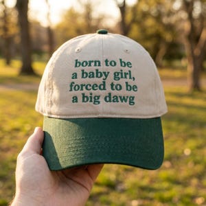 May include: A two-toned baseball cap with a beige crown and a dark green brim. The front of the crown features dark green embroidered text that reads: born to be a baby girl, forced to be a big dawg. The cap has a button on the top center.