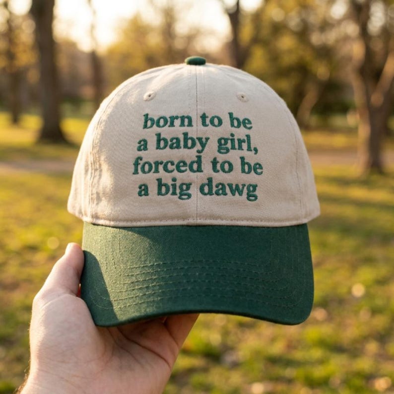 May include: A two-toned baseball cap with a beige crown and a dark green brim. The front of the crown features dark green embroidered text that reads: born to be a baby girl, forced to be a big dawg. The cap has a button on the top center.