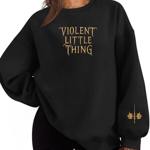 May include: Black sweatshirt with the text "VIOLENT LITTLE THING" in gold lettering across the chest. A gold emblem is on the left sleeve. The sweatshirt has long sleeves and a crew neck.