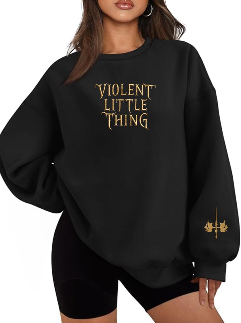 May include: Black sweatshirt with the text "VIOLENT LITTLE THING" in gold lettering across the chest. A gold emblem is on the left sleeve. The sweatshirt has long sleeves and a crew neck.