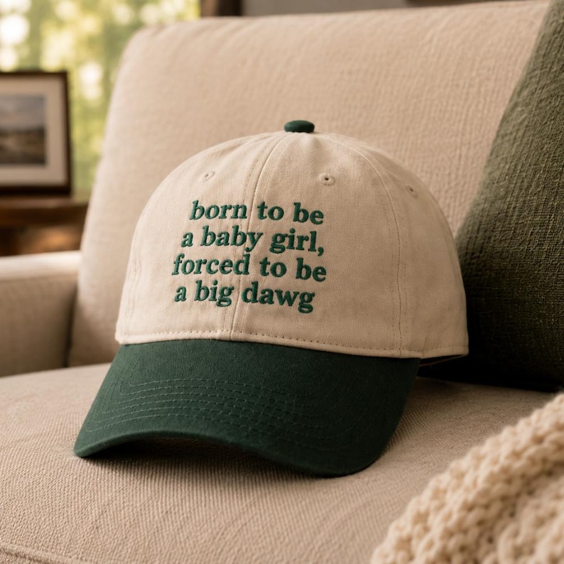 May include: A beige baseball cap with a dark green brim and a dark green embroidered text on the front that reads: born to be a baby girl, forced to be a big dawg. The cap features a button on the top and stitched eyelets for ventilation.
