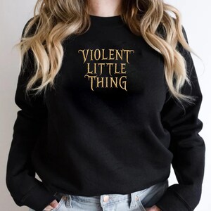 May include: Black crew neck sweatshirt with the text "VIOLENT LITTLE THING" in a gold, stylized font. The sweatshirt is a solid black color and has long sleeves. The text is centered on the chest.