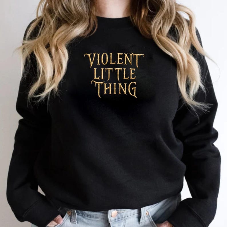 May include: Black crew neck sweatshirt with the text "VIOLENT LITTLE THING" in a gold, stylized font. The sweatshirt is a solid black color and has long sleeves. The text is centered on the chest.