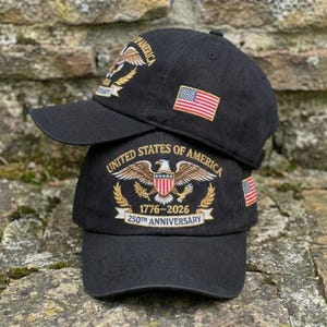 Puede incluir: Dos gorras de b&eacute;isbol negras apiladas una encima de la otra. Cada gorra presenta un dise&ntilde;o bordado con las palabras "United States of America" y un &aacute;guila. Una gorra muestra la bandera estadounidense. La gorra inferior dice "1776-2026 250th Anniversary."