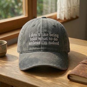 May include: Gray baseball cap with the embroidered text "I don't like being told what to do unless I'm naked." The hat is made of a soft, worn fabric and has a curved brim. The hat is sitting on a wooden surface.