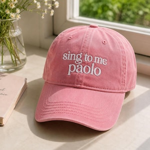 May include: A dusty pink baseball cap featuring the white embroidered text sing to me paolo on the front panel. The cap has a structured crown, a curved brim with visible stitching, and a button at the top center of the six-panel construction.