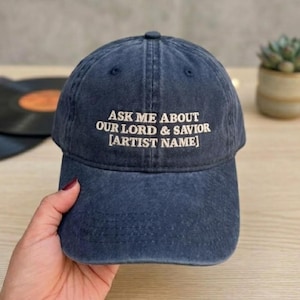 May include: A dark blue baseball cap with a washed-out, distressed texture. The front features white embroidered text that reads: ASK ME ABOUT OUR LORD & SAVIOR [ARTIST NAME]. The cap has a curved brim and a structured crown with a top button.