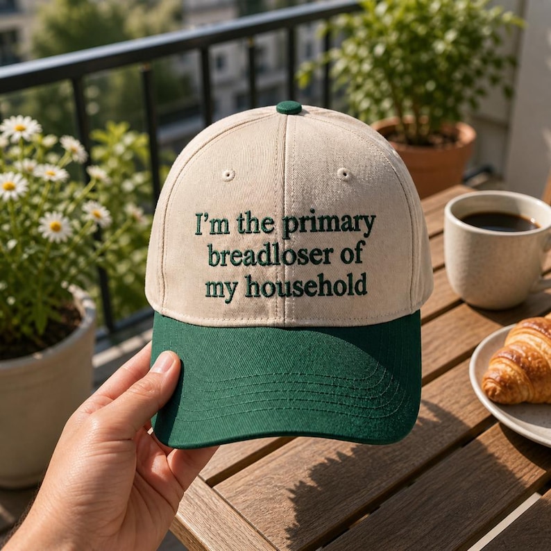 May include: A beige and green baseball cap with the embroidered text, "I'm the primary breadloser of my household." The cap has a green brim and a green button on top. The hat is held by a person.