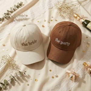 May include: Two baseball caps, one beige and one brown, are displayed on a cream-colored surface. The beige cap reads "the bride" in a matching beige thread, while the brown cap says "the party" in white thread. The caps are surrounded by decorative elements.