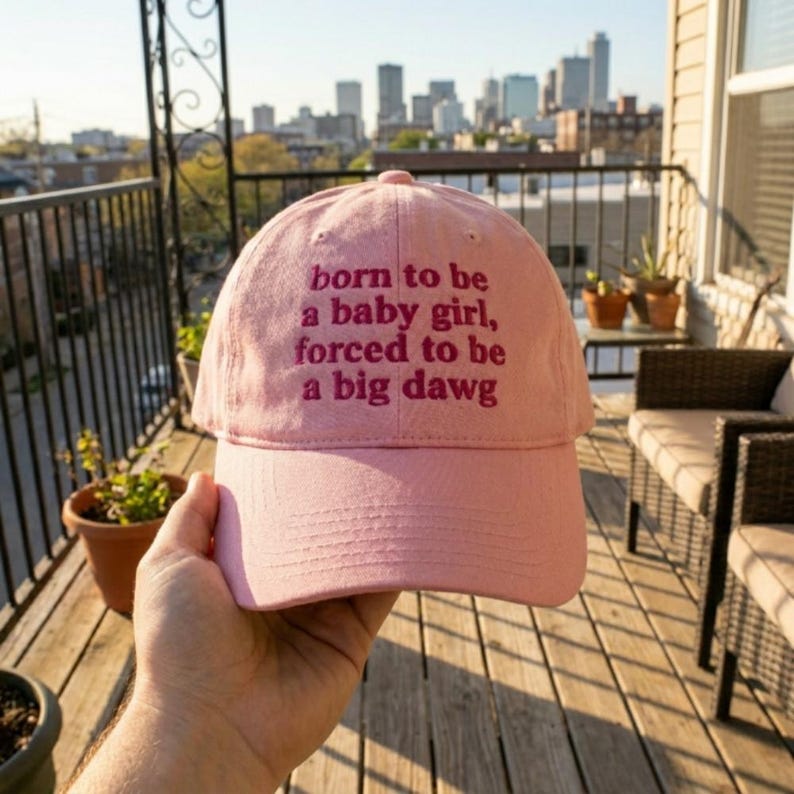 May include: A light pink baseball cap featuring the embroidered text "born to be a baby girl, forced to be a big dawg" in a dark pink font. The cap has a structured crown, a curved brim, and a classic six-panel design with stitched eyelets.