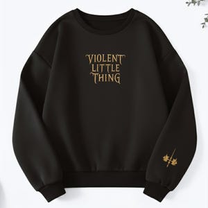 May include: Black sweatshirt with the text "VIOLENT LITTLE THING" in gold script across the chest. A gold graphic of a stylized weapon is on the left sleeve. The sweatshirt has a crew neck and ribbed cuffs and hem.