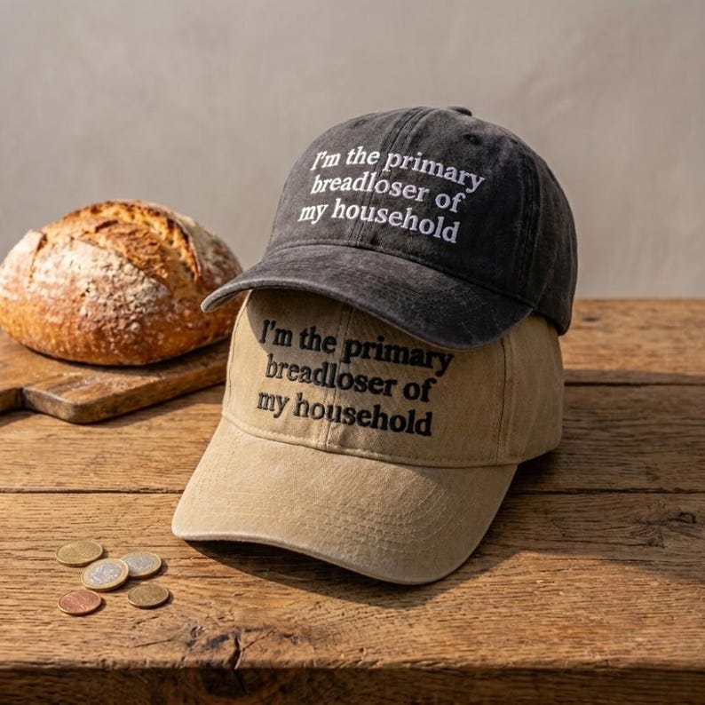 May include: Two baseball caps, one black and one tan, stacked on top of each other. Each cap has the embroidered text "I'm the primary breadloser of my household". A loaf of bread and some coins are in the background.