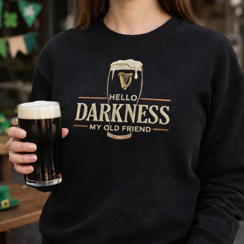 May include: Black sweatshirt with the phrase "Hello Darkness My Old Friend" in gold lettering. The design includes a pint of stout with a harp symbol. A person is holding a pint of stout.