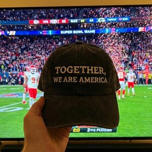 K&ouml;nnte beinhalten: Dunkelgraue Baseballkappe mit dem wei&szlig;en, gestickten Text "TOGETHER, WE ARE AMERICA". Die Kappe wird vor einem Fernsehbildschirm gehalten, der ein Footballspiel zeigt.