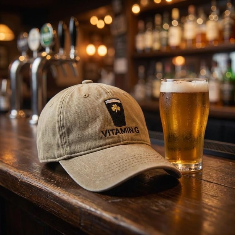 K&ouml;nnte beinhalten: Khaki-Baseballkappe mit dem Wort "VITAMING" unter einer Grafik eines Bierkrugs mit einem Kleeblatt. Ein Glas Bier steht neben der Kappe auf einer Holzbar. Der Hintergrund zeigt eine Bar mit Zapfh&auml;hnen und Flaschen.