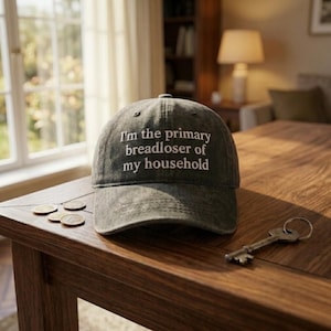 May include: A gray baseball cap with the text "I'm the primary breadloser of my household" embroidered in white. The cap sits on a wooden table with coins and a key.
